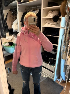 Ljusrosa hoodie från Peak Performance - Säljer en ljusrosa hoodie från Peak Performance med dragkedja, huva och vita dragsnören. Hoodien har en broderad logga på bröstet och ett märke på ärmen. Mjuk och skön bomullsblandning, perfekt för chill dagar.
