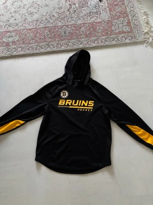 Boston Bruins träningströja  - En officiell Boston Bruins träningshoodie med emblem och lagnamn samt NHL loggan på ena arm från Fanatics och deras authentic pro modell. Längst ner i armarna är insydda några cm på ett snyggt sätt då armarna var lite långa. Vill man fixa tillbaka de kan stygnen tas bort efter betalning och beställning. Storlek L 