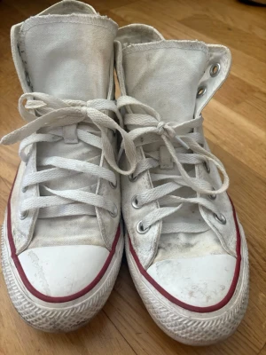 Vita Converse Chuck Taylor Highs - Klassiska vita Converse Chuck Taylor All Star high-tops med röd och blå rand runt sulan. Skorna är i canvas med vita snören och metallöljetter. Ikonisk logga på insidan av skaftet. Perfekta för en avslappnad och cool stil.kommer att självklart tvättas innan skick 
