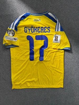 Svensk landslagströja Gyökeres #17 - Gul och blå svensk landslagströja från Adidas med Gyökeres och nummer 17 på ryggen.FIFA och UEFA-märken på ärmarna samt klassiska Adidas-ränder på axlarna. Tillverkad i lätt och ventilerande material. Adidas plast påse ingår också i köpet! samt  kan priset diskuteras!