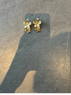 Blommiga örhängen i guld och vitt - Säljer ett par örhängen med blommotiv i guld och vitt. Örhängena har flera små blommor i metall och detaljer som ger en elegant och unik look. Perfekta för dig som vill ha något som sticker ut och känns trendigt.