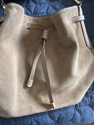 Beige mocka bucket bag från Ralph Lauren - Snygg använd beige bucket bag i mocka från Ralph Lauren med justerbar axelrem och dragskostängning. Perfekt för dig som gillar en stilren och trendig look. Priset reflekterar skicket på väskan. Finns en fläck som säkert går att ta bort.