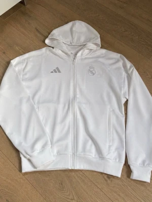 Vit Real Madrid hoodie från Adidas - Snygg vit hoodie med dragkedja från Adidas och Real Madrid-logga på bröstet. Klassisk huva och två fickor framtill. Perfekt för dig som gillar sportig stil och vill visa ditt stöd för laget.