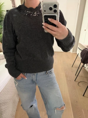Mörkgrå stickad tröja med paljetter - Mysig mörkgrå stickad tröja med rund hals och dekorativa paljetter runt kragen. Tröjan har långa ärmar och ribbade muddar vid ärmslut och nederkant. Perfekt för dig som vill ha något extra till vardagslooken.