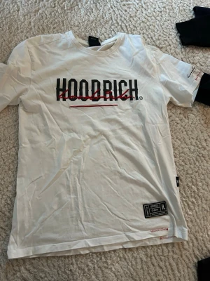 Vit HOODRICH t-shirt med tryck - Säljer en vit t-shirt från HOODRICH i storlek M. T-shirten har ett stort svart och rött tryck på bröstet och en patch nertill. Klassisk rund hals och korta ärmar. Passar även S