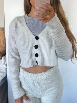 Kofta - Säljer en vit croppad kofta med ribbad struktur och fyra stora svarta knappar framtill. Koftan har v-ringning och långa ärmar, perfekt för en chill och trendig look. Råkat tvätta den med mörka kläder därav går den mot det grå hållet. Storlek S 🩷