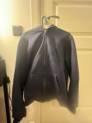 Mörkblå hoodie från Polo Ralph Lauren - Mörkblå hoodie med dragkedja från Polo Ralph Lauren. Klassisk design med huva, fickor framtill och det ikoniska röda Polo-logot broderat på bröstet. Skön bomullskänsla och perfekt för en avslappnad look.