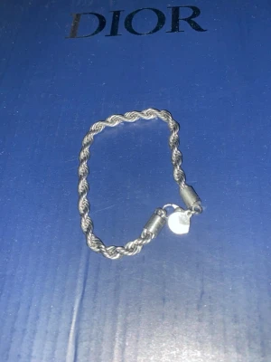 Silverfärgat armband från Dior - Snyggt silverfärgat armband från Dior med tvinnad kedja och en liten rund berlock. Armbandet har ett stilrent och elegant utseende, perfekt för att lyfta din outfit. Materialet är metall och designen är klassisk med en modern twist.