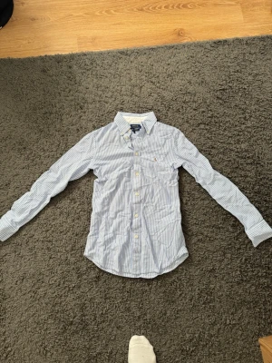 Blårandig skjorta från Polo Ralph Lauren - Klassisk blårandig skjorta från Polo Ralph Lauren med broderad logga på bröstet. Skjortan har lång ärm, knappar framtill och är gjord i mjuk bomull. Perfekt för en stilren och avslappnad look. Lite mindre i storlek 