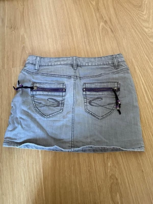 Grå kort jeanskjol med dragkedjor - Snygg grå kort jeanskjol med två bakfickor som har lila dragkedjor och dekorativa snören. Storlek 32, men passar även 34! 