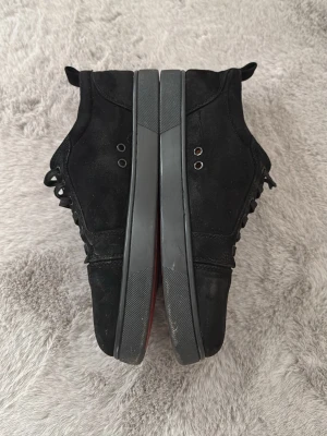 Svarta Christian Louboutin sneakers - Snygga svarta sneakers från Christian Louboutin med klassisk röd sula och svarta mockadetaljer. Skorna har svarta snören, metallöljetter och en stilren design som sticker ut med den ikoniska röda undersidan.