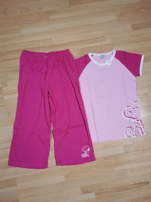 Snoopy rosa pyjamas set - Supergulligt tvådelat pyjamas set från Snoopy i rosa och ljusrosa bomull. T-shirten har raglanärmar och tryck med Snoopy, byxorna är vida och har resår i midjan samt en liten Snoopy-detalj. Perfekt för mysiga kvällar hemma!