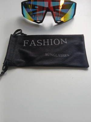 Färgglada sportiga solglasögon - Snygga sportiga solglasögon med spegelglas i flera färger som blått, rött och gult. Modellen har en kantig, futuristisk form och kommer med en svart tygpåse med texten 'FASHION SUNGLASSES'. Perfekta för dig som vill sticka ut i sommar. Oanvända och har bra kvalité.