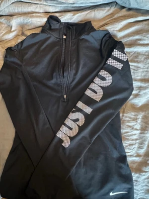 Svart träningströja Nike Just Do It - Svart långärmad träningströja från Nike med halv dragkedja framtill och hög krage. På ena ärmen står det 'JUST DO IT' i stora vita bokstäver. Materialet är stretchigt och andas, perfekt för löpning eller gym.