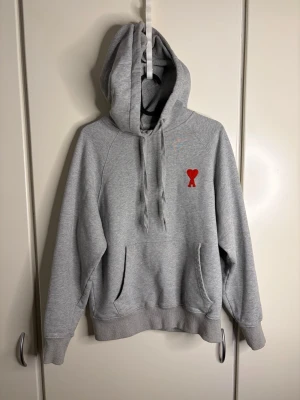 Grå hoodie från Ami Paris - Snygg grå hoodie från Ami Paris med röd hjärtlogga på bröstet. Klassisk passform med stor huva, dragsko och känguruficka. Tillverkad i mjuk bomullsblandning som känns skön mot huden. Perfekt för en avslappnad och trendig look.