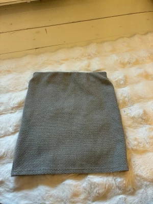 Grå kortkjol i strukturerat tyg - Snygg grå kortkjol med strukturerat vävt tyg och rak passform. Kjolen är enkel och stilren, perfekt att matcha med olika toppar för en trendig look.