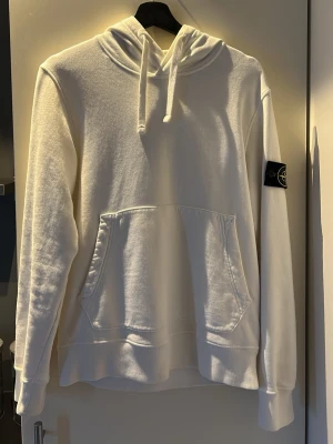 Vit hoodie från Stone Island - Säljer en stilren vit hoodie från Stone Island med klassisk huva och känguruficka framtill. Tröjan har den ikoniska loggan på vänster ärm och är gjord i mjukt material som känns skönt mot huden. Perfekt för dig som gillar streetwear och enkel design.