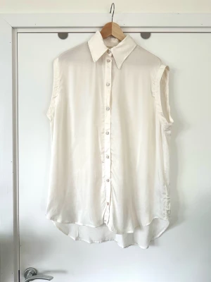 Ärmlös vit blus i silkeslook - H&M storlek S - Stilren helt NY vit ärmlös blus / skjorta / topp från H&M i storlek S. Tidlös modell med med klassisk krage och knappar framtill. Skjortan har en behagligt lös passform och är tillverkad i mjukt, silkeslent tyg i 100% viskos, så bra material som andas. Helt ny med etikett!