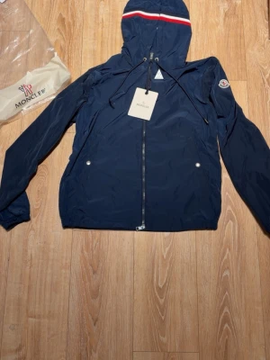 Marinblå vindjacka från Moncler - Snygg marinblå vindjacka från Moncler med huva och dragkedja framtill. Jackan har Moncler-logga på ärmen och en rödvit rand på huvan. Lätt material och två fickor med tryckknappar. Perfekt för blåsiga dagar.