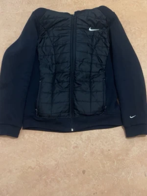 Svart hoodie från Nike med quiltad front - Snygg svart hoodie från Nike med quiltad frontpanel och dragkedja. Hoodien har två fickor med dragkedja framtill och Nike-logga på bröstet och ärmen. Tillverkad i mjukt material med en sportig look och huva baktill.