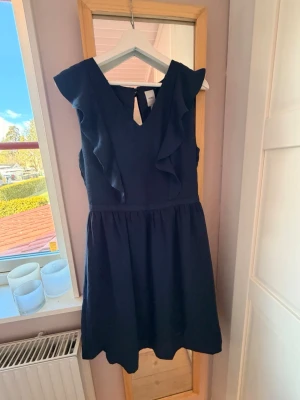 Mörkblå volangklänning med v-ringning - Supersöt mörkblå klänning med v-ringning och volangdetaljer längs axlarna. Klänningen är ärmlös och har en lätt utställd kjol som ger en feminin siluett. Perfekt för dig som gillar stilrena och eleganta plagg.