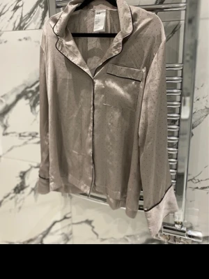 Satin pyjamas - H&M - Snygg tvådelad pyjamas i beige satin med små prickar och svarta kantband. Skjortan har krage, bröstficka och knäppning framtill, byxorna har resår i midjan. Inga skador eller defekter 