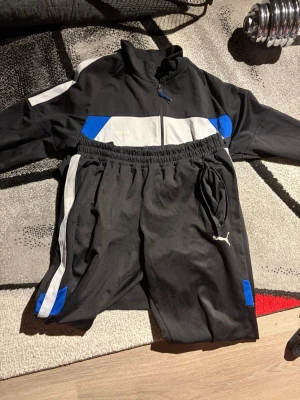 Svart loungewear-set från Puma - Säljer ett svart loungewear-set från Puma med vita och blå detaljer. Setet består av en zip-jacka med hög krage och matchande byxor med resår och dragsko i midjan. Materialet är mjukt och bekvämt, perfekt för att chilla hemma. Puma-loggan finns på både jacka och byxor.