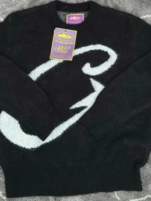 Svart stickad tröja från CORTEIZ - Cool svart stickad tröja från CORTEIZ med ett stort vitt grafiskt mönster på framsidan. Tröjan har rund halsringning och ribbstickade muddar vid ärmslut och nederkant. Perfekt för dig som gillar unik design och mjuka material.