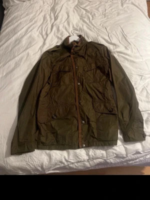 Olivgrön vindjacka med fickor - Snygg olivgrön vindjacka med fyra stora fickor framtill och knäppning med både dragkedja och knappar. Jackan har hög krage och är tillverkad i ett lätt, vattenavvisande material. Perfekt för blåsiga dagar och enkel att matcha med olika outfits.