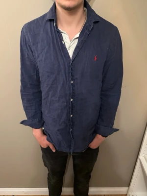 Marinblå skjorta från Ralph Lauren - Säljer en marinblå skjorta från Ralph Lauren med klassisk krage och knappar framtill. Skjortan har lång ärm och den ikoniska röda loggan broderad på bröstet. Tillverkad i mjuk bomull som känns skön mot huden. Alldrig använd