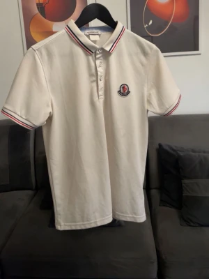 Vit Moncler pikétröja - Klassisk snygg vit pikétröja från Moncler med röd och marinblå rand på krage och ärmslut. Tröjan har korta ärmar, knappslå framtill och Moncler-logga broderad på bröstet. Perfekt nu till vår och sommar och passar mycket bra till jeans och shorts.