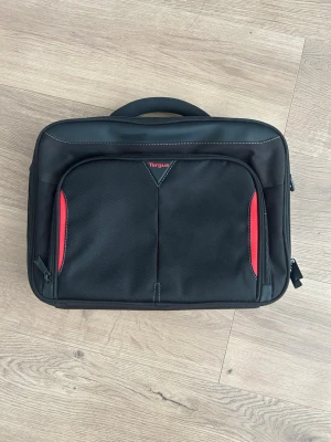 Svart datorväska från Targus - Snygg svart datorväska från Targus med röda detaljer på framsidan. Väskan har ett stort huvudfack, praktiskt bärhandtag och extra ytterfack med dragkedja. Tillverkad i slitstarkt syntetmaterial och har en stilren, rektangulär form.