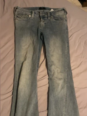 Ljusblå bootcut jeans True Religion - Säljer ett par ljusblå bootcut jeans från True Religion med klassiska fickor bak och snygga slitningar framtill. Jeansen har låg midja och är tillverkade i denim med stretch för extra komfort. Perfekta för en avslappnad och trendig look.