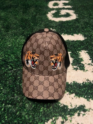 GG keps med tigertryck - Snygg keps från GG i beige med klassiskt GG-mönster och två tigrar broderade framtill. Baksidan är i svart mesh för extra ventilation och kepsen har böjd skärm. Perfekt för dig som vill sticka ut med en exklusiv accessoar.