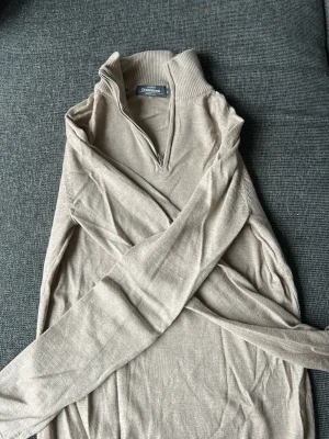 Beige merinoulltröja från Dressmann - Stickad beige tröja från Dressmann i mjuk merinoull. Tröjan har halv dragkedja framtill, hög krage och långa ärmar. Perfekt för lager-på-lager och enkel att matcha med olika outfits.