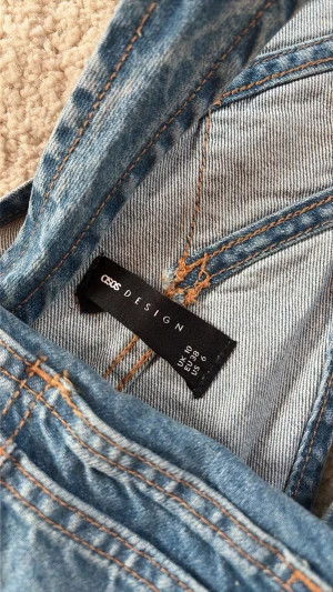 Blå denim hängselbyxor ASOS DESIGN - Säljer ett par klassiska blå denim hängselbyxor från ASOS DESIGN. Byxorna har justerbara axelband, stor bröstficka och flera knappar i sidorna. Perfekt för en avslappnad och trendig look. Materialet är 100% bomull med kontrastsömmar.