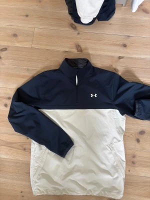 Under Armour vindjacka - Snygg vindjacka från Under Armour i marinblått och vitt. Jackan har halv dragkedja, hög krage och två sidofickor. Lätt material som skyddar mot vind, perfekt för aktiva dagar. Under Armour-logga på bröstet ger en sportig look.