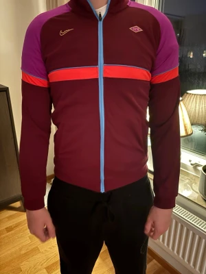 Nike sportjacka vinröd/lila - Snygg sportjacka från Nike i vinrött med lila axelpartier, röda och blå detaljer samt hel dragkedja framtill. Jackan har lång ärm och är tillverkad i ett stretchigt funktionsmaterial, perfekt för träning eller uppvärmning.