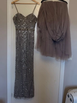 Glittrig långklänning med tyllkjol - Säljer en elegant långklänning i silver med tunna axelband och hjärtformad urringning. Klänningen är täckt av glittriga paljetter och har en avtagbar tyllkjol i grått som ger extra volym. Perfekt för bal eller festliga tillfällen.