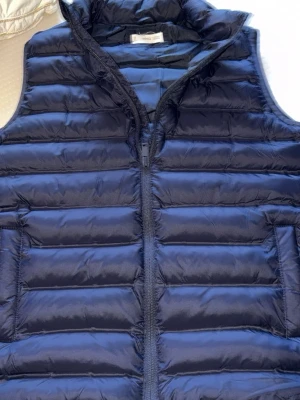 Mörkblå dunväst - Snygg mörkblå dunväst med hög krage och quiltad design. Västen har dragkedja framtill och två sidofickor. Perfekt att slänga över en hoodie eller tröja för extra värme. Tillverkad i lätt och smidigt material med dunfyllning.