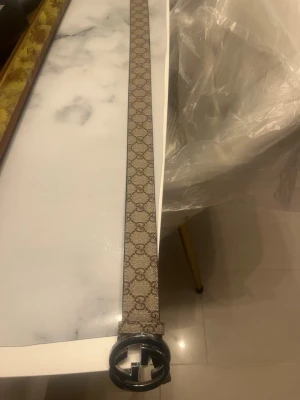 Beige GG-bälte från Gucci - Snyggt beige bälte från Gucci med klassiskt GG-mönster och rund metallspänne med GG-logga. Bältet är gjort i canvas med läderdetaljer och har en stilren design som passar till många outfits.