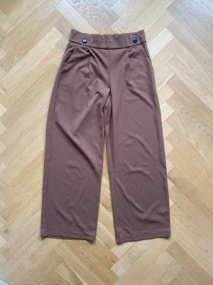 Ljusbruna vida byxor med knappar - Säljer ett par ljusbruna byxor med två svarta dekorativa knappar i midjan. Materialet känns mjukt och har ett snyggt fall.. Clean och stilren look.