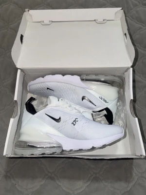 Nike Air Max 270 vita sneakers - Nike Air Max 270 sneakers i vitt med svart swoosh och detaljer. Ovandel i mesh för bra ventilation, synlig Air-enhet i hälen och klassisk snörning. Snygg och sportig design som passar till det mesta. Perfekta för dig som gillar stilrena och bekväma sneakers.