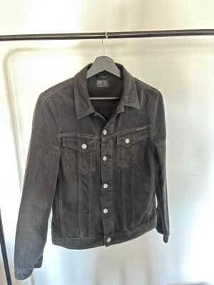Svart jeansjacka från Nudie Jeans  - Snygg svart jeansjacka från Nudie Jenas med klassisk krage, silverfärgade knappar och två bröstfickor med lock. Jackan har raka ärmar och justerbara knappar i midjan. Perfekt för en cool och avslappnad stil.