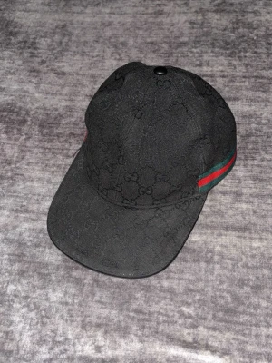 Svart Gucci keps med GG-mönster - Säljer en svart keps från Gucci med det klassiska GG-monogrammönstret  över hela kepsen. På sidan finns en grön och röd rand som ger en ikonisk touch. Kepsen har böjd skärm och justerbar rem bak. Materialet känns slitstarkt och exklusivt.