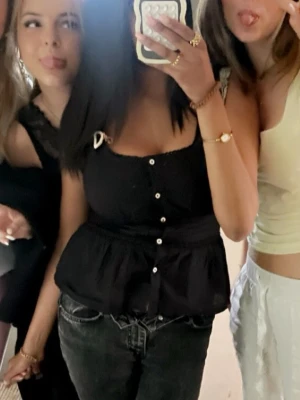 Svart blus med knappar och volang - Säljer en svart blus med knappar framtill och volang nedtill. Toppen har breda axelband, varav ett med rosettdetalj, och en feminin passform. 