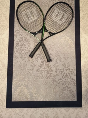Två svarta tennisracketar Wilson - Säljer två snygga tennisracketar från Wilson i svart med gröna och vita detaljer. Racketarna har klassisk form och greppvänliga handtag. Perfekt för dig som vill spela tennis med stil och bra känsla.