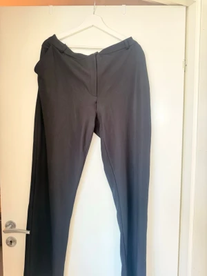 Svarta kostymbyxor med raka ben - Snygga svarta kostymbyxor med klassisk rak passform och hällor för skärp. Byxorna har en enkel design med dold dragkedja och knapp framtill. Perfekta för en stilren look. Materialet känns mjukt och följsamt.