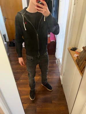 Svart hoodie från Ralph Lauren - Svart zip-hoodie från Ralph Lauren med vit dragsko i huvan och röd broderad logga på bröstet. Klassisk modell med fickor framtill och mjukt bomullsmaterial. Perfekt för en avslappnad och stilren look. 