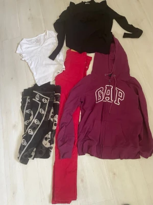 Klädpaket med hoodie, tröjor och byxor - Klädpaket med vinröd GAP-hoodie med dragkedja, svart långärmad tröja, vit t-shirt, röda byxor och svarta leggings med dödskallemönster. Perfekt för dig som gillar en mix av basic och edgy stil. Flera färger och mönster för att variera din look.